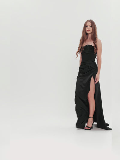 Model prezentând o rochie elegantă din satin negru cu corset și deschidere laterală, capturând fluiditatea materialului și silueta sofisticată.